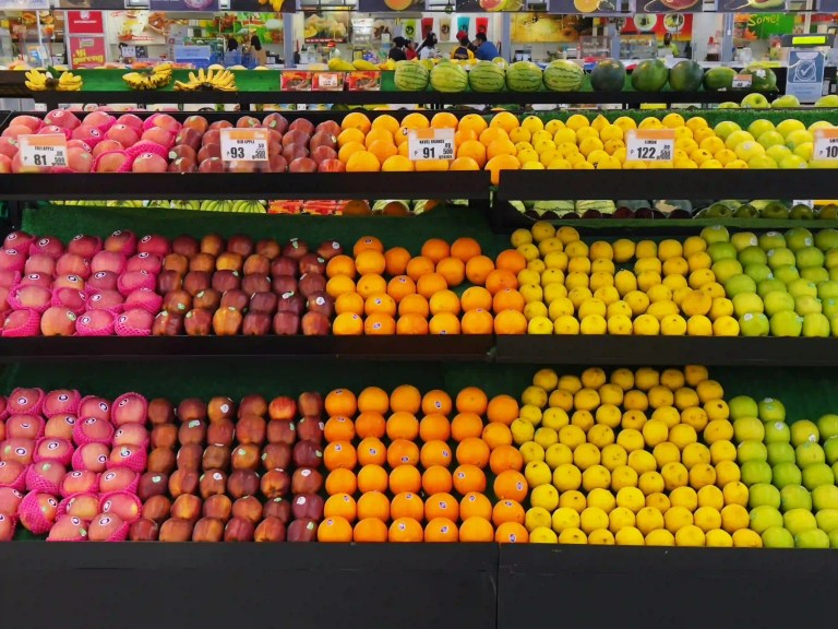 A neatly arranged supermarket fruit display featuring rows of colorful fruits, including pink apples, red apples, oranges, lemons, and green apples. The fruits are organized by color in bright, symmetrical sections, creating a rainbow-like gradient across the shelves.