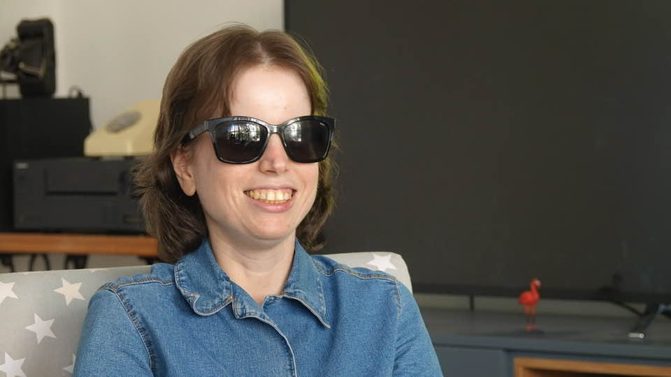 Tali sitting in a chair wearing dark sunglasses and smiling widely.
