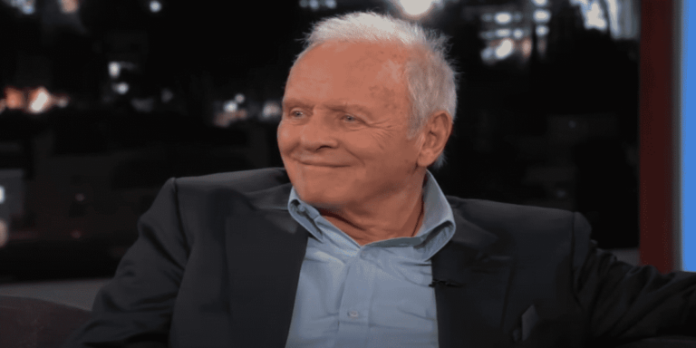 Sir Anthony Hopkins on a TV show, wearing a blue shirt and dark-colored jacket. He's sitting facing the audience and smiling.