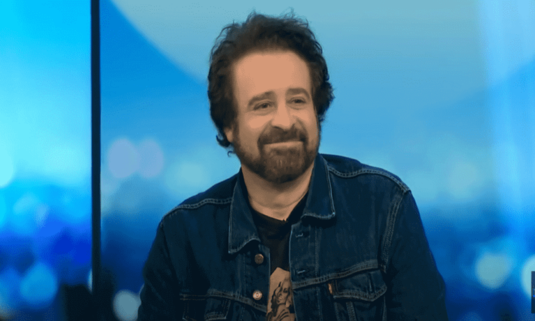 Adam Duritz on a live talk show. He is a middle-aged man with short curly hair and close-cropped beard. He's wearing a t-shirt and a denim jacket.