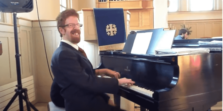 Ben Monkaba playing on a piano, smiling broadly. He is a young red-haired man wearing glasses and a suit.