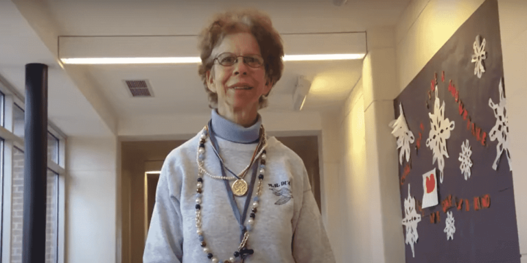 Gloria Lenhoff standing in a white passageway. She wearing an off-white sweater over a turtleneck shirt, horn-rimmed glasses and several neck pieces including a rosary and a medal.