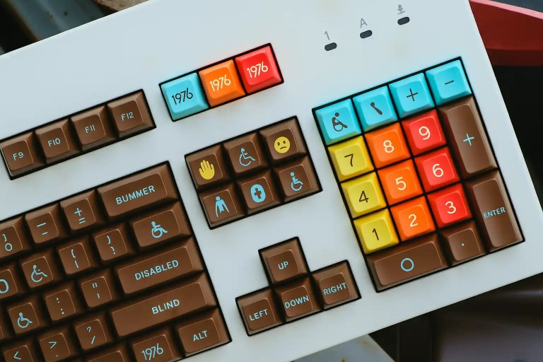 A colorful mechanical keyboard featuring brown, blue, yellow, and orange keycaps. Several keys display accessibility icons, including symbols for wheelchair access, visual impairment, sign language, and assistance. The design blends a retro aesthetic with a modern focus on inclusion and accessibility.